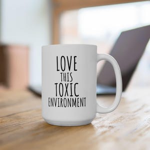 Puede incluir: Taza de cerámica blanca con el texto "LOVE THIS TOXIC ENVIRONMENT" en negro, fuente sans-serif. La taza tiene un asa curva y está colocada sobre una superficie de madera, con un portátil en el fondo.