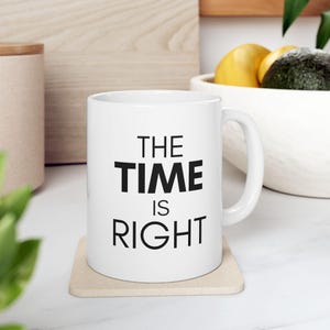 Könnte beinhalten: Weiße Keramik-Tasse mit dem Text "THE TIME IS RIGHT" in Schwarz. Die Tasse steht auf einem beigen Untersetzer. Ein Holzbehälter und eine Schüssel mit Obst sind im Hintergrund.