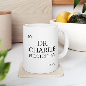 May include: A white ceramic mug with the text "It's DR. CHARLIE ELECTRICIAN to you" in black. The mug is on a beige coaster. The background includes a wooden surface and a bowl with fruit.