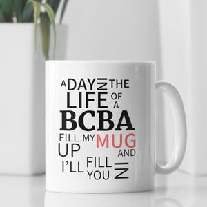 May include: White ceramic mug with black text that reads "A Day in the Life of a BCBA Fill My Mug Up and I'll Fill You In". The word "Mug" is in pink.