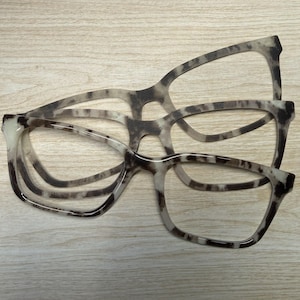 May include: Three pairs of eyeglasses with a speckled pattern in shades of brown and cream. The frames have a rectangular shape with rounded edges. The glasses are displayed on a light-colored wooden surface.