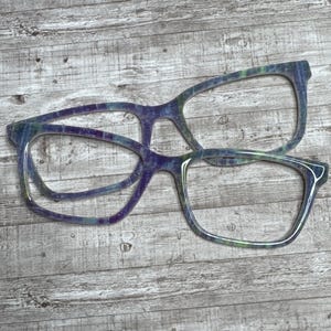 May include: Two pairs of glasses with a tie-dye pattern. The glasses are layered on top of each other. The glasses are made of a clear material and have a purple, blue, and green tie-dye pattern.