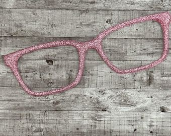 May include: A pink glitter eyeglasses frame prop. The glasses are a classic rectangular shape, with a thick frame. The prop is made of a sparkly, glittery material. The background is a weathered, gray wooden surface.
