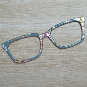 May include: A pair of eyeglasses with a floral pattern. The frames are a muted teal color, adorned with a delicate design featuring white, pink, and yellow flowers. The glasses are set against a light wood grain background.