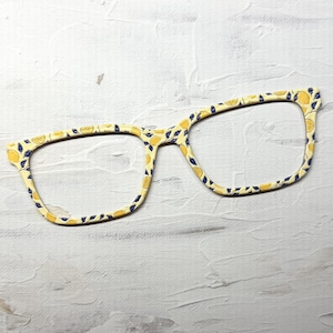 May include: A pair of eyeglass frames featuring a light yellow base color with a repeating pattern of small yellow lemons and dark blue leaves. The frames have a rectangular shape with rounded corners and a thin, lightweight construction.