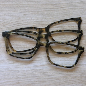 May include: Two pairs of eyeglasses with a brown and black tortoiseshell pattern frame. The frames feature a rectangular shape with rounded edges and are constructed from a smooth, polished material. The glasses are stacked slightly offset.