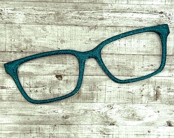 May include: Teal-colored, glitter-covered eyeglasses frame. The frame is in a classic rectangular shape, with a sparkly finish. The glasses are set against a light-colored, wooden background.