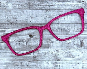 May include: A pair of bright pink, glitter-covered eyeglasses. The glasses have a rectangular frame shape and are set against a light gray and white wood-grain background. The glasses are a solid color with no lenses.