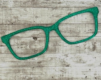 May include: A pair of oversized, glittery green eyeglasses. The frames are a bright, solid green color and have a thick, bold outline. The glasses are a decorative item, likely for a party or photo booth.