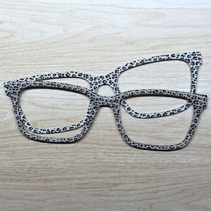 May include: Two flat, two-dimensional eyeglass frames featuring a repeating black and grey leopard print pattern. The frames are shaped with rectangular lenses and are positioned overlapping each other, showcasing the animal print design clearly.
