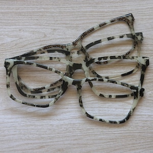 May include: Two pairs of eyeglasses frames stacked on top of each other. The top pair features a translucent frame with gold glitter accents. The bottom pair has a black and white tortoiseshell pattern with a square-shaped frame design.