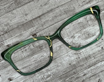 May include: Green cat-eye glasses with gold accents on a wood background.