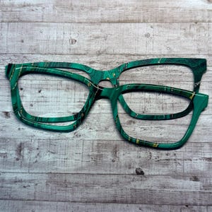 May include: Two pairs of eyeglasses with a malachite pattern. The frames are a vibrant green with gold accents, creating a marbled effect. The glasses are set against a light gray wooden background.