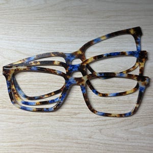 May include: Two pairs of eyeglass frames stacked on top of each other. The frames feature a mottled pattern with a blend of brown, tan, and bright blue colors. The frames have a rectangular shape with rounded edges and a smooth, glossy finish.