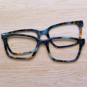 May include: Two pairs of eyeglasses with a marbled design in shades of blue, gold, and purple. The frames have a rectangular shape and are set against a light wood grain background. The glasses are empty of lenses.