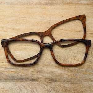 May include: Two pairs of eyeglasses with a wood grain pattern. The frames are brown and black, with a rectangular shape. One pair is stacked on top of the other, with the lenses removed. The background is a light brown wooden surface.