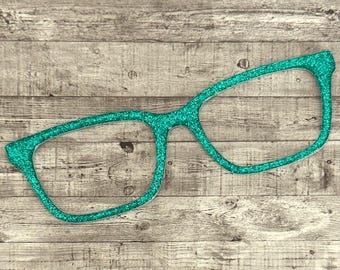 May include: A teal glitter eyeglasses cutout. The glasses have a classic rectangular frame shape. The cutout is placed on a wooden background.