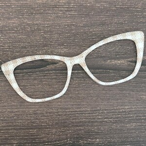 May include: A decorative pair of eyeglasses with a white and tan plaid pattern. The glasses are cut out and have a retro cat-eye shape. The background is a dark brown wood grain.