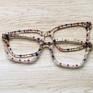 May include: Clear eyeglasses with a red, white, and blue star pattern. The frames have a rectangular shape and are decorated with small stars. The glasses are set against a light wood grain background.