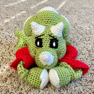 May include: A green crocheted triceratops dinosaur toy with white horns and eyes. The dinosaur is wearing a red crocheted scarf.