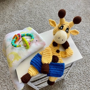 May include: A crocheted giraffe toy with brown spots and blue denim shorts. The giraffe has a yellow body, brown ears, and a white face with black eyes. The toy is lying on a white surface.