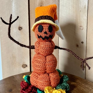 Può includere: Una figura a forma di zucca realizzata a uncinetto con un cappello e un viso amichevole. La figura ha braccia di filo marrone ed è circondata da fiori all'uncinetto in rosso, verde e giallo.