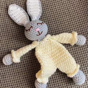 Peut inclure: Un lapin en peluche crocheté avec des oreilles grises et un corps crème. Le lapin a un visage souriant avec des joues roses et les yeux fermés.