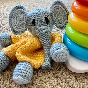 May include: A crocheted elephant toy with a yellow dress. The elephant has blue ears and a gray trunk. The toy is sitting on a white surface.