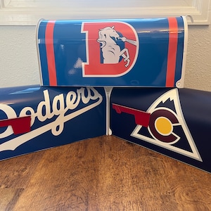 May include: Three blue mailboxes with different sports team logos. The top mailbox has a red and white Denver Broncos logo, the middle mailbox has a white Los Angeles Dodgers logo, and the bottom mailbox has a white Colorado Avalanche logo.