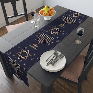May include: A navy blue table runner featuring gold menorahs, Star of David symbols, and the text "Happy Hanukkah." The runner is on a dark wooden table, set with plates, cutlery, and wine glasses. A bowl of apples is in the centre.
