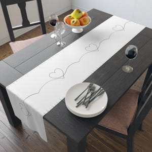 May include: A white table runner with a black heart design, set on a dark wooden table. The table is set with plates, forks, wine glasses, and a bowl of apples. The runner adds a decorative touch to the dining setting.