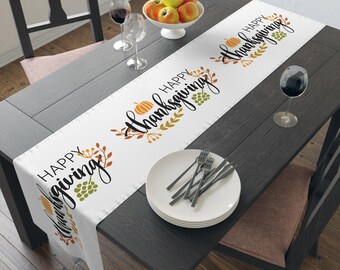 May include: A white table runner with the words "Happy Thanksgiving" in black script, featuring orange pumpkins and green grape illustrations. The runner is on a dark wooden table, set with plates, cutlery, and wine glasses.