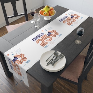 Puede incluir: Camino de mesa blanco con un diseño patriótico que presenta el texto "AMERICA 250" en rojo y azul, junto con gráficos de estrellas y fuegos artificiales. El camino de mesa está colocado sobre una mesa de madera oscura, con un cuenco de fruta y copas de vino.