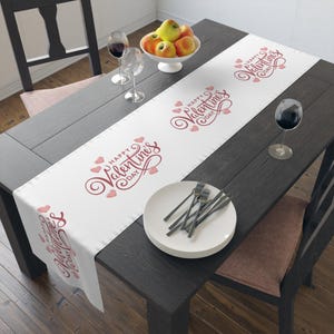 Puede incluir: Un camino de mesa blanco de San Valentín con las palabras "Happy Valentine's Day" en escritura roja y acentos de corazón. El camino de mesa está sobre una mesa de madera oscura, con platos, tenedores y copas de vino.