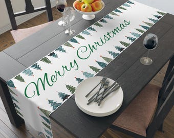 Puede incluir: Camino de mesa navideño blanco con texto verde "Merry Christmas". El camino de mesa está decorado con árboles de Navidad verdes y azules. Está colocado sobre una mesa de madera oscura con platos, cubiertos y copas de vino.