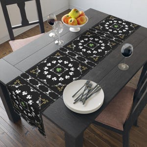 May include: A black table runner with a repeating pattern of white shamrocks and gold accents. The runner features a central shamrock design in green. The table is set with plates, silverware, and wine glasses, with a bowl of fruit.