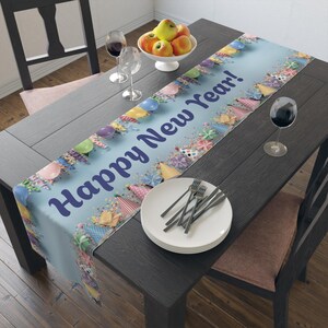 Peut inclure: Une nappe de table bleu clair avec le texte "Happy New Year !" en bleu foncé. La nappe est décorée de ballons colorés, de chapeaux de fête et de confettis. Une assiette blanche avec des fourchettes et un bol de pommes sont sur la table.