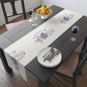 May include: A white table runner with the text "Happy Mother's Day" in purple script and a purple flower design. The runner is on a dark wooden table set with plates, silverware, and wine glasses. A bowl of apples sits in the center.