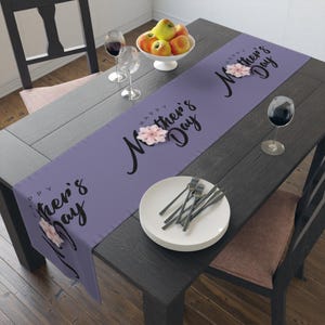 May include: A lavender table runner with the text "Happy Mother's Day" in black script and a pink flower design. The runner is on a dark wooden table set with plates, silverware, and wine glasses. A bowl of apples sits in the background.