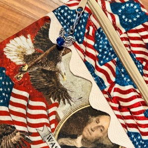 May include: A zippered pouch with a patriotic design, featuring American flags, eagles, and a portrait of George Washington. The pouch has a beige zip and a decorative angel charm with a blue bead. The fabric is red, white, and blue.