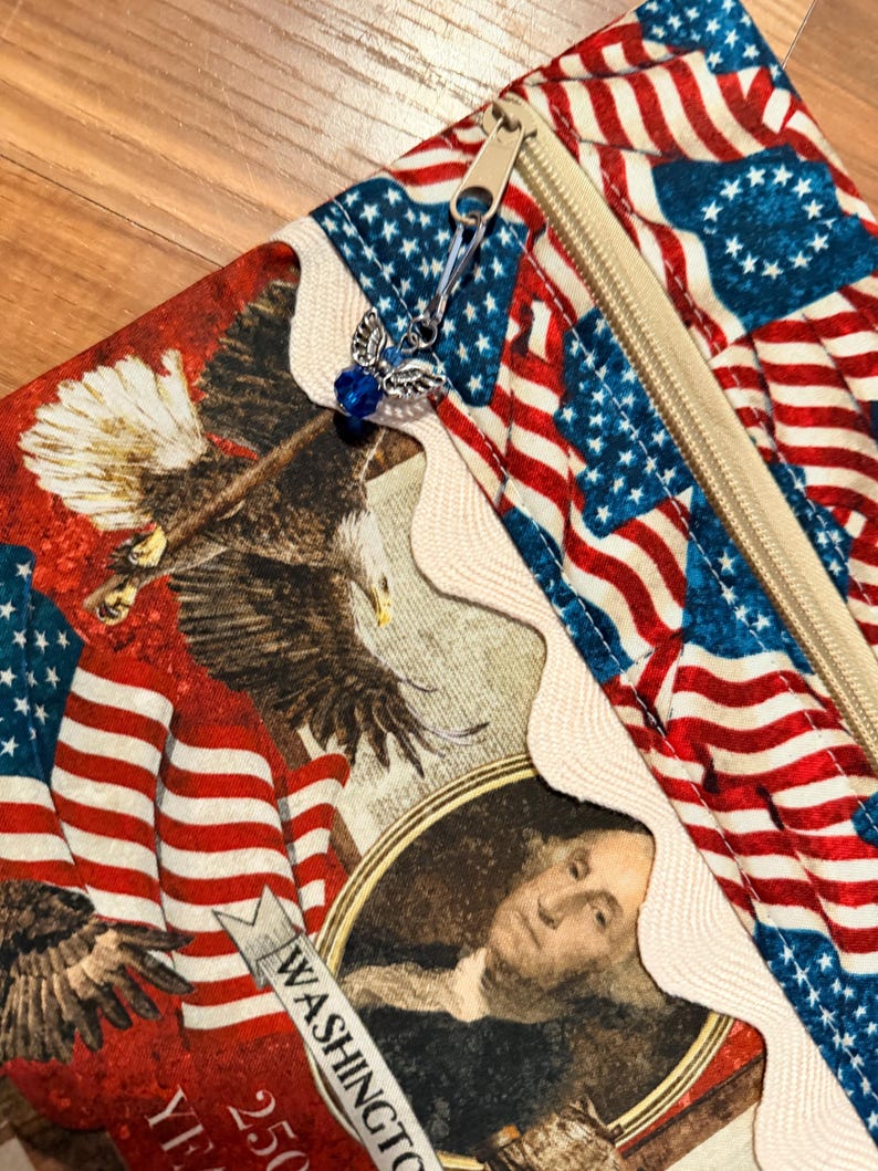 May include: A zippered pouch with a patriotic design, featuring American flags, eagles, and a portrait of George Washington. The pouch has a beige zip and a decorative angel charm with a blue bead. The fabric is red, white, and blue.