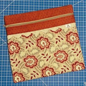 Puede incluir: Una bolsa rectangular con cremallera y un estampado floral. La bolsa presenta un fondo beige con grandes flores rojas y rosas y hojas verdes. La sección superior es de color óxido sólido con cierre de cremallera. Un ribete de encaje festoneado separa las dos secciones.