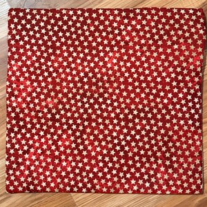 May include: A red fabric with a repeating pattern of white stars. The fabric is rectangular and appears to be folded. The stars are evenly distributed across the surface. The fabric is lying on a wooden surface.