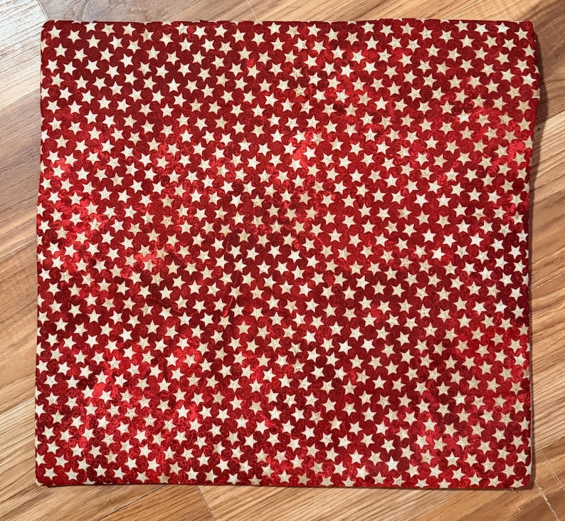 May include: A red fabric with a repeating pattern of white stars. The fabric is rectangular and appears to be folded. The stars are evenly distributed across the surface. The fabric is lying on a wooden surface.