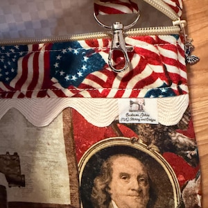 May include: A zippered pouch with a patriotic American flag design. The pouch features a red, white, and blue colour scheme with a zip closure and a silver-coloured metal clasp. The design includes a portrait of Benjamin Franklin.