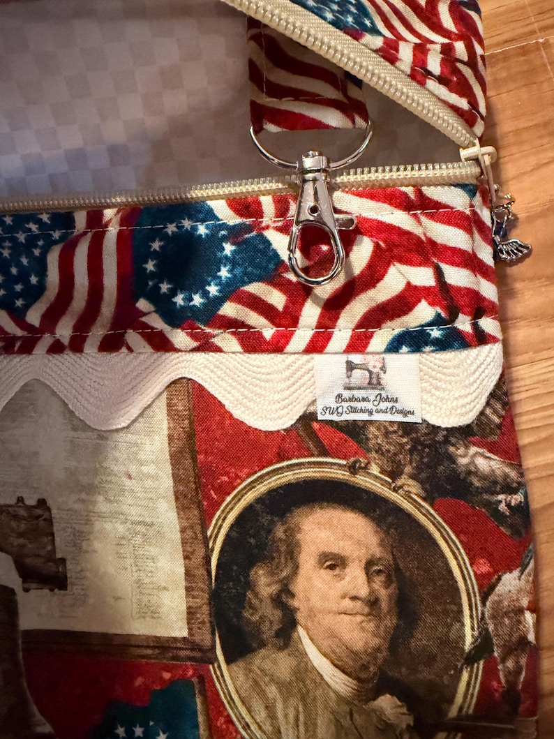 May include: A zippered pouch with a patriotic American flag design. The pouch features a red, white, and blue colour scheme with a zip closure and a silver-coloured metal clasp. The design includes a portrait of Benjamin Franklin.