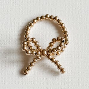 May include: A gold-toned beaded ring with a bow design. The ring is made of small, round beads that are connected together to form a circle. The bow is created by twisting the beads together in the center of the ring.