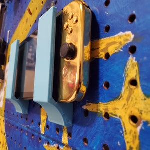 Puede incluir: Dos soportes de pared de plástico azul claro para una consola de videojuegos, con una consola Nintendo Switch dorada montada en el lado derecho.