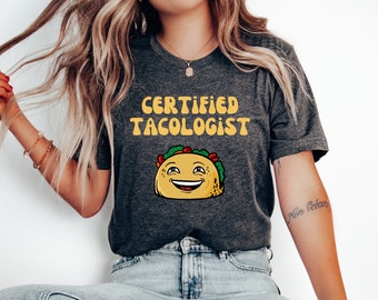 May include: Dark gray t-shirt with the text "CERTIFIED TACOLOGIST" in yellow, above a smiling cartoon taco. The taco has a yellow shell, green lettuce, and red tomato. A fun, food-themed graphic tee.