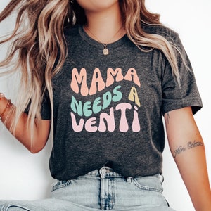 May include: A dark grey t-shirt with the text "Mama Needs a Venti" in colorful, retro-style lettering.